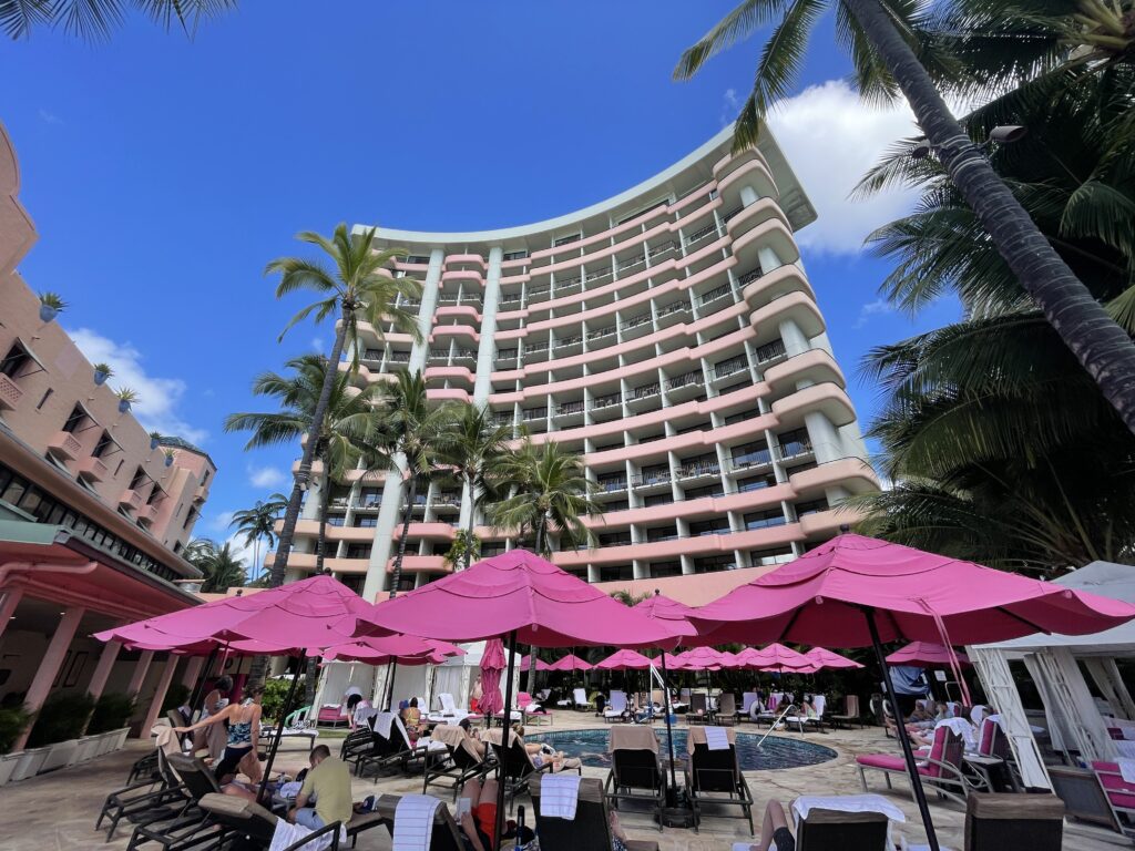 5 Key Features To Know When Deciding Between The Sheraton Waikiki Vs. Royal Hawaiian Hotel Hawaii Calling a hotel with pink umbrellas and palm trees