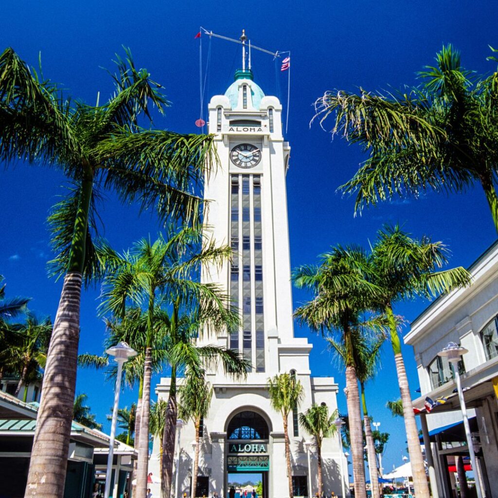 Oahu Tour Crowned Number One on TripAdvisor's Global 2023 Best of the Best Hawaii Calling a tall white tower with a clock on top and palm trees with Aloha Tower in the background